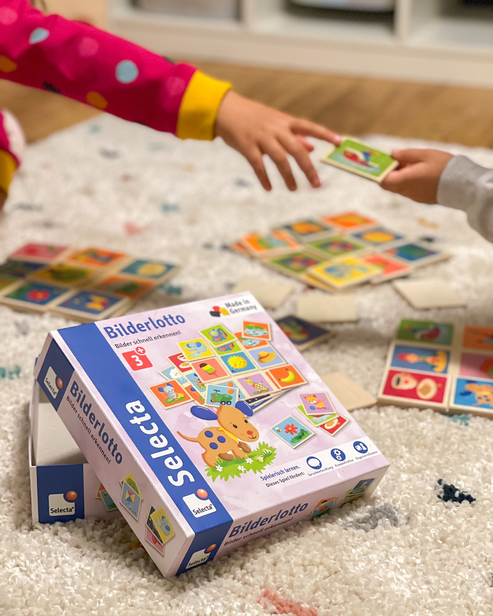 Im Vordergrund liegt die geöffnete Kartonverpackung des Selecta Bilderlotto. Dahinter liegen auf einem beigefarbenen Teppichboden die Spielkarten und Plättchen. an erahnt zwei Kinderhände, die sich gerade eine Bilderkarte reichen.