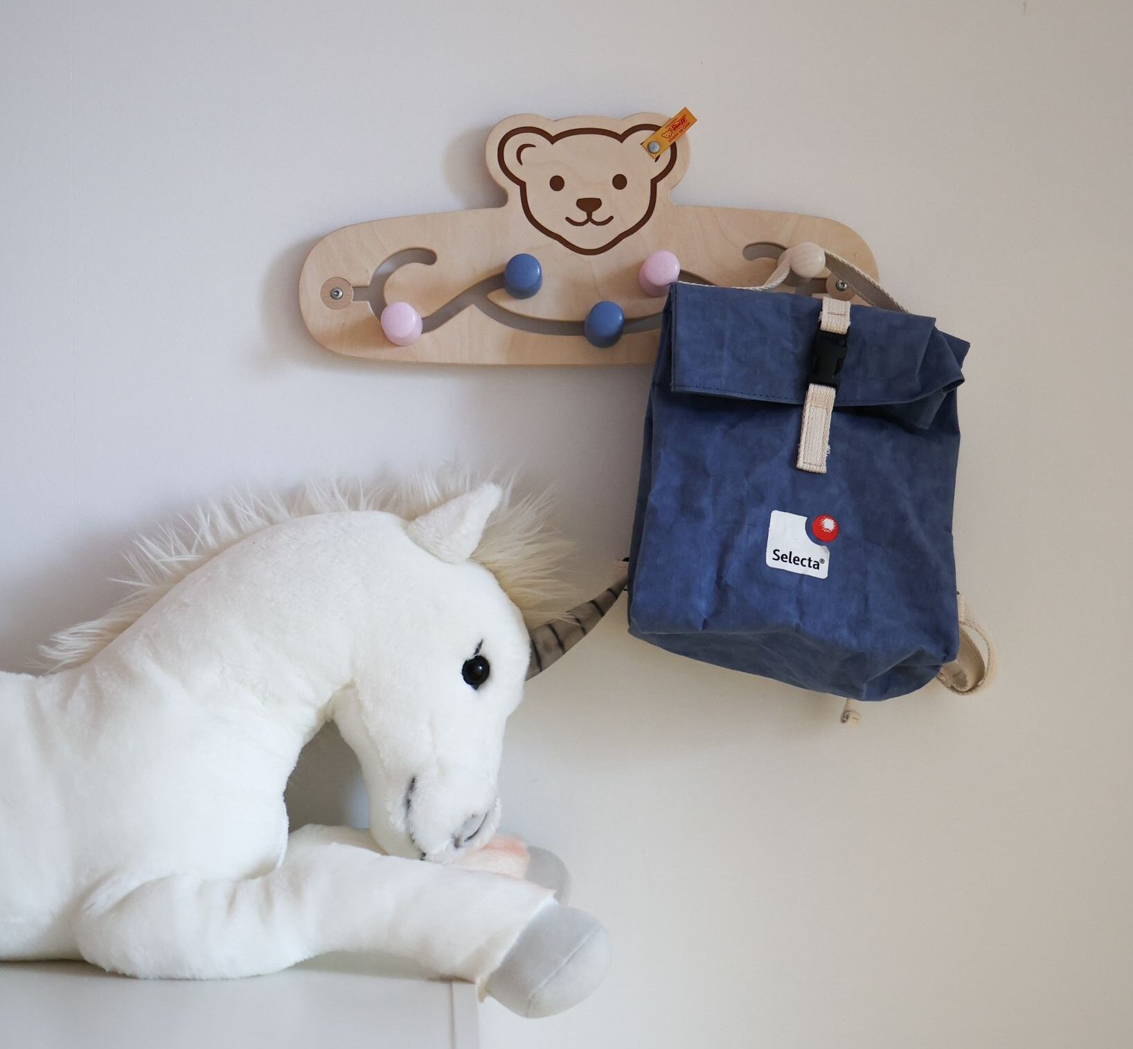 Eine Kindergarderobe aus Holz von Steiff hängt an einer weißen Wand. Die Aufhängelemente sind von links nach rechts rosa, blau, blau, rosa und holzfarben. Am ganz rechten Element ist ein dunkelblauer Rucksack aufgehängt, der die Aufschrift Selecta trägt. Im Bild links unten sieht man ein weißes Plüscheinhorn, dass auf einem Schrank liegt.